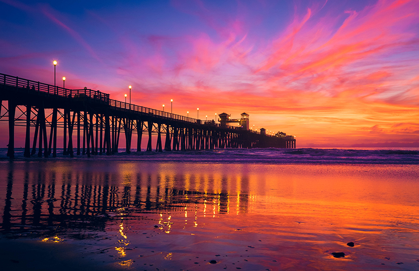 Colorful sunset over a wooden fishing pier with reflections across the shoreline at low tide.
